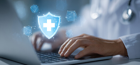 A healthcare professional types on a laptop, with a glowing shield icon featuring a medical cross above, symbolizing digital health security.