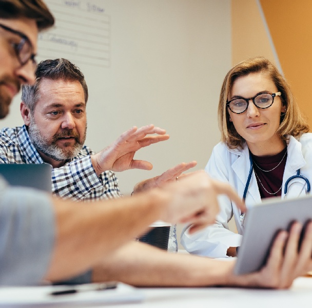A group of professionals engages in a discussion around a table, collaborating with a tablet and notes in a bright, modern meeting space.