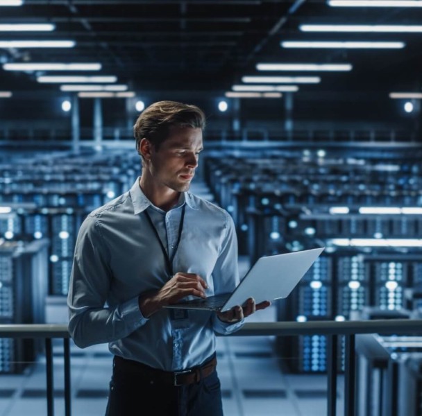 A person in a blue shirt stands in a high-tech data center, typing on a laptop with rows of servers illuminated in the background.