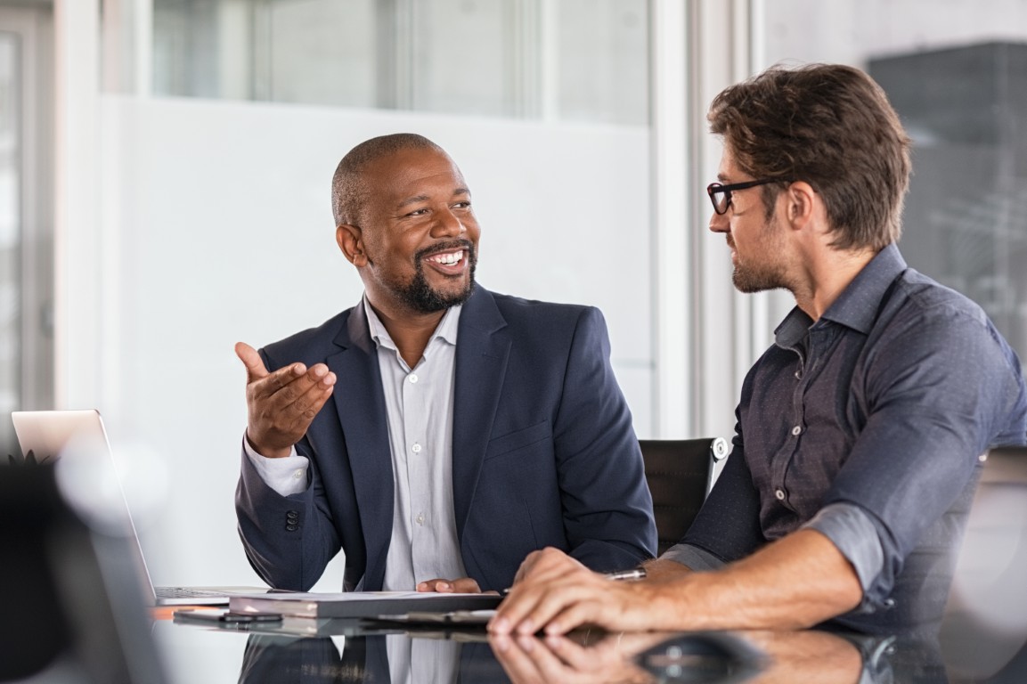 Two professionals engaged in a discussion at a modern conference table, with laptops and documents in view.