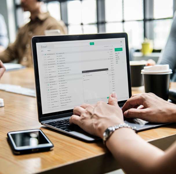A person types on a laptop displaying an email interface, surrounded by colleagues and coffee on a wooden table.