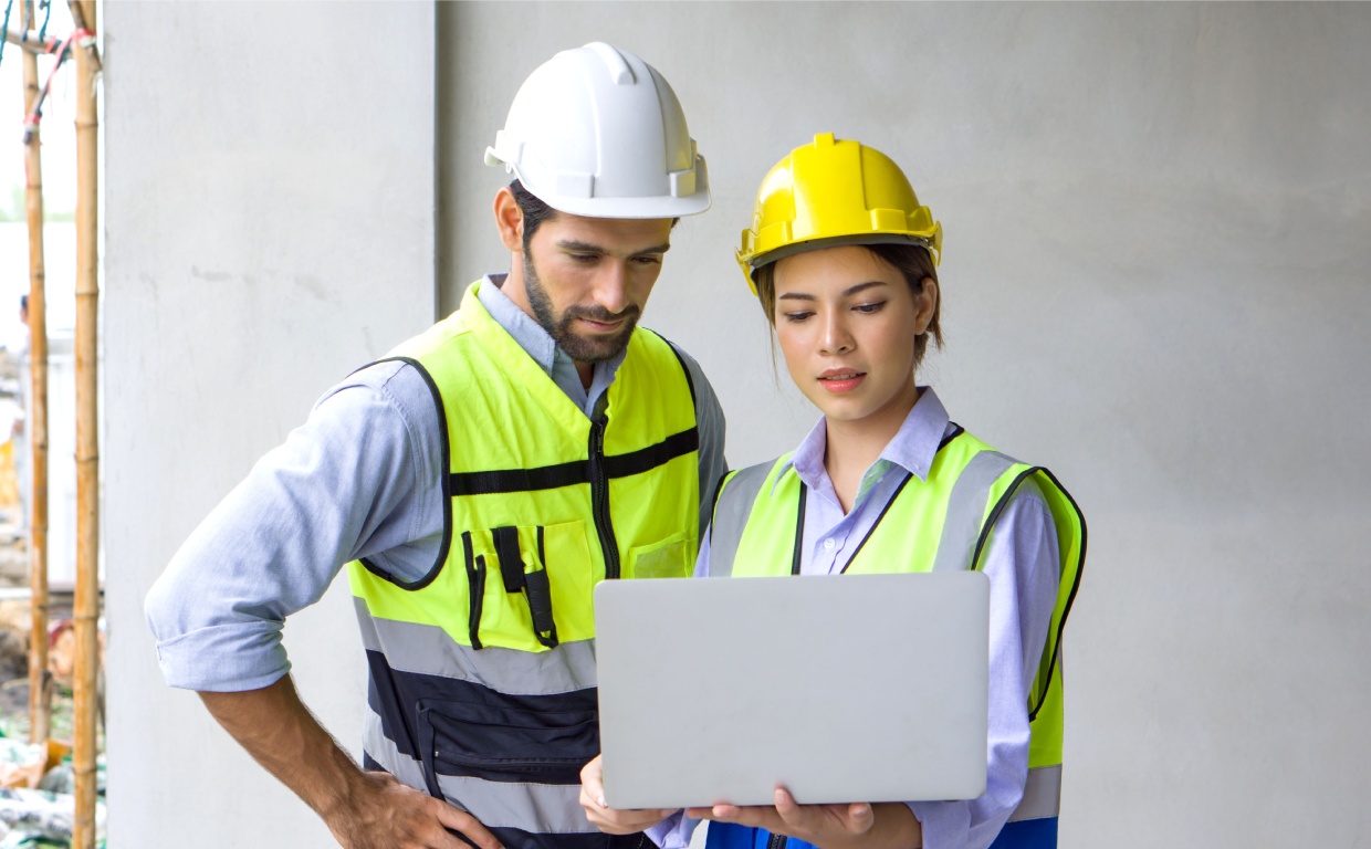 Two construction workers, one in a white hard hat and the other in a yellow hard hat, review plans on a laptop in a building site.