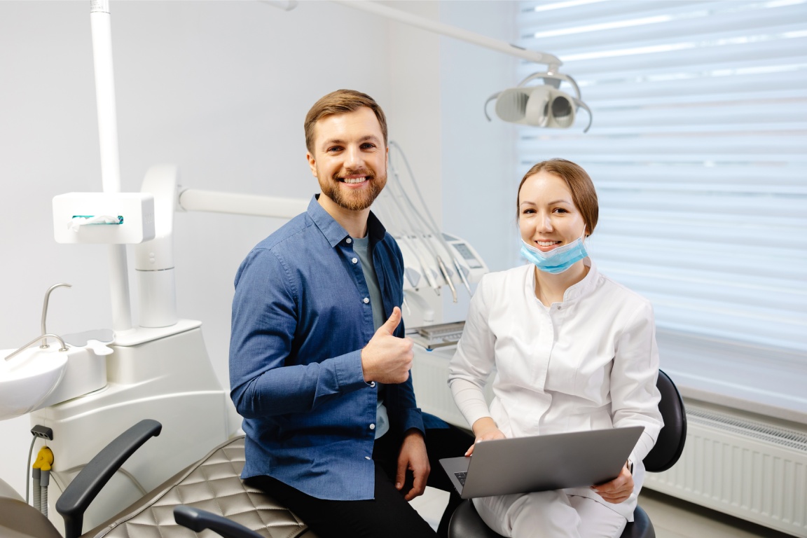 A person in a blue shirt gives a thumbs-up while sitting next to a dental professional with a laptop in a modern dental office.