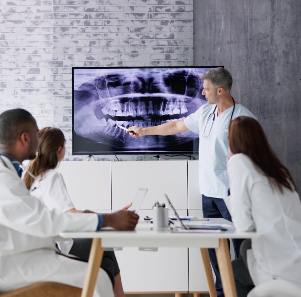 A group of medical professionals observes a dental X-ray presentation, as a doctor points out details on a screen.