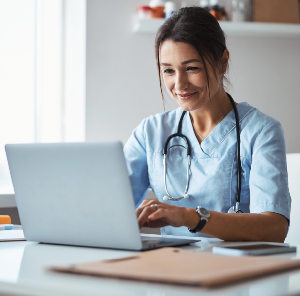 A healthcare professional in scrubs works on a laptop in a bright, modern office, with a stethoscope around their neck.
