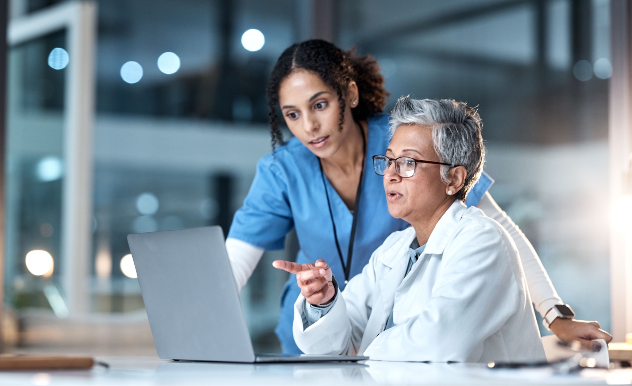 Two healthcare professionals are engaged in a conversation while reviewing information on a laptop in a modern medical setting.