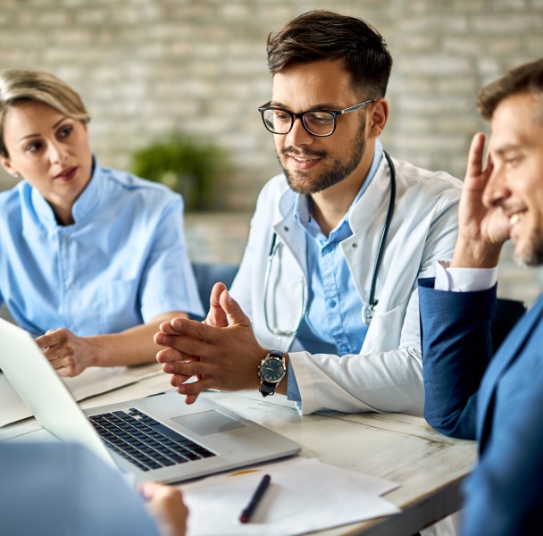 A group of medical professionals and a businessman discuss over a laptop in a bright, modern setting.