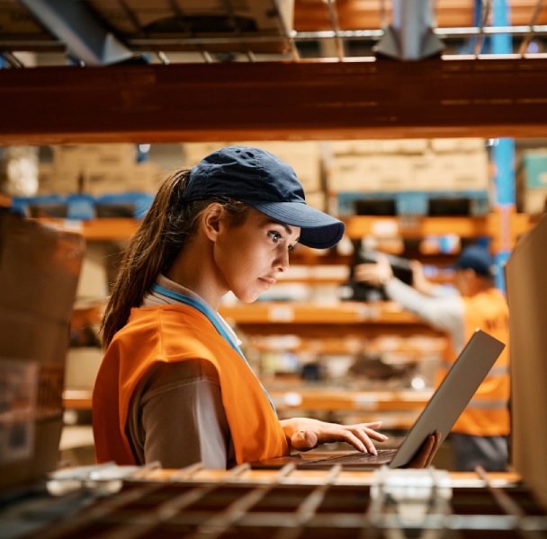 A worker in an orange vest and cap uses a tablet in a warehouse filled with boxes and shelves, while another employee organizes items in the background.