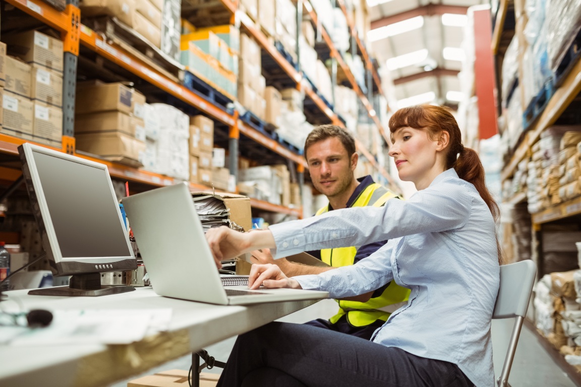 Two workers collaborate at a desk in a warehouse, using a laptop amidst stacks of boxes and storage shelves.