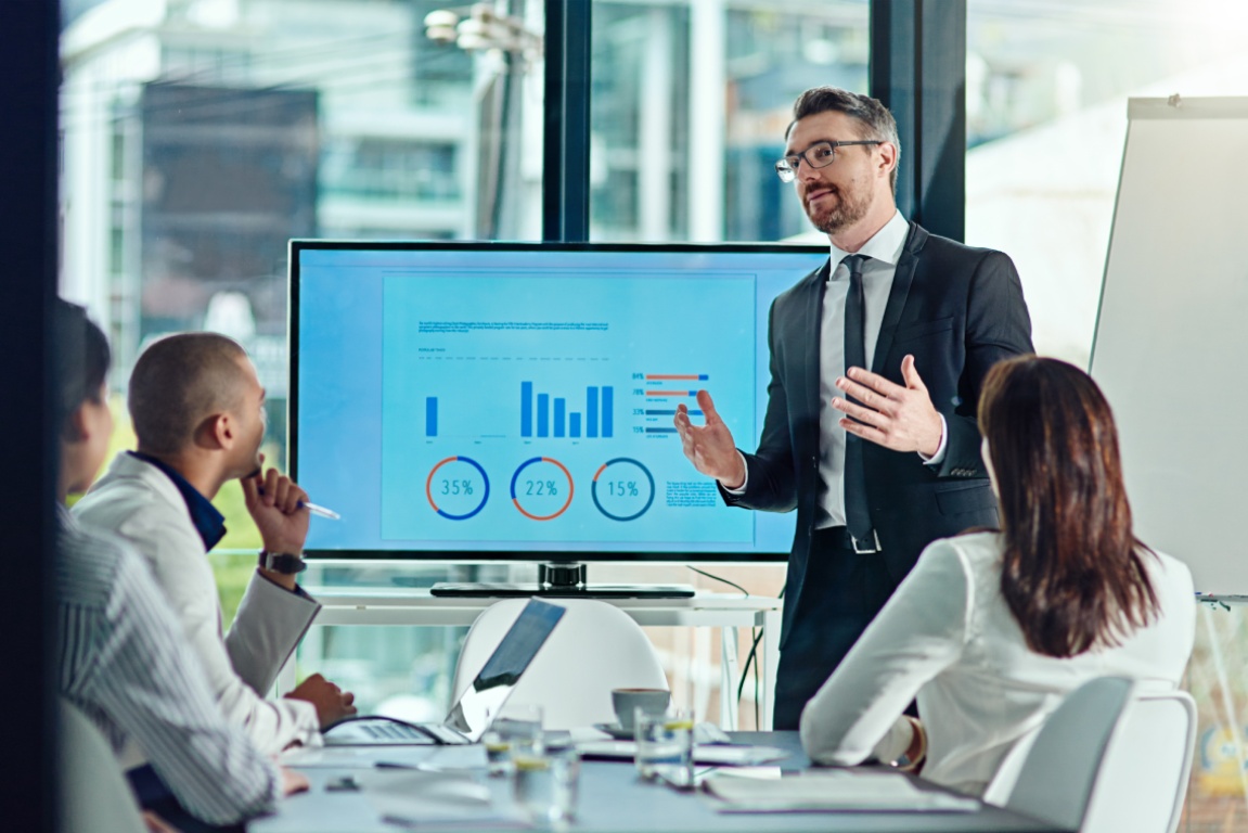 A businessman presents data on a screen during a boardroom meeting, with colleagues engaged in discussion around the table.