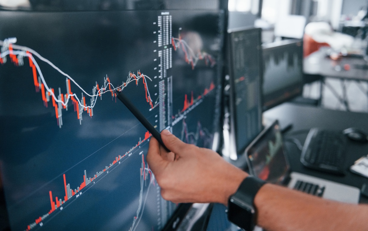 A hand pointing at a colorful financial graph on a large screen, displaying market trends and data analysis in a modern office setting.