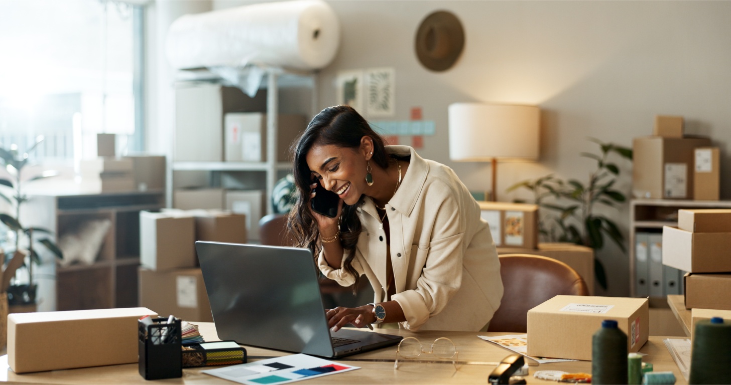 A person is on the phone while working on a laptop in a cozy workspace filled with cardboard boxes and plants.