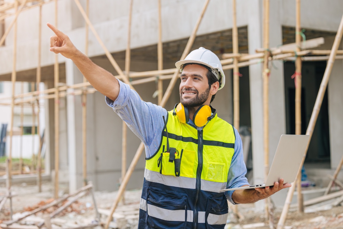 A construction worker in a safety vest and hard hat points upwards while holding a laptop on a building site.