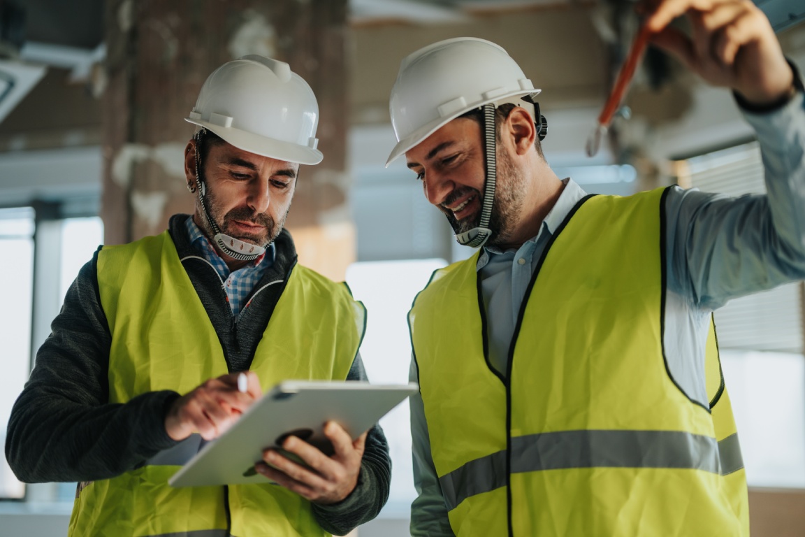Two construction workers in hard hats and high-visibility vests, one holding a tablet and the other raising a tool.