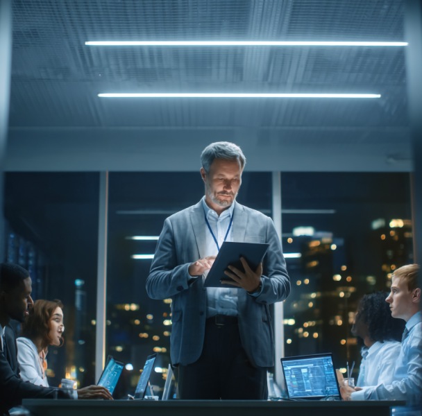 A business meeting in a modern office with a leader presenting on a tablet, while team members work on laptops in a city skyline setting.