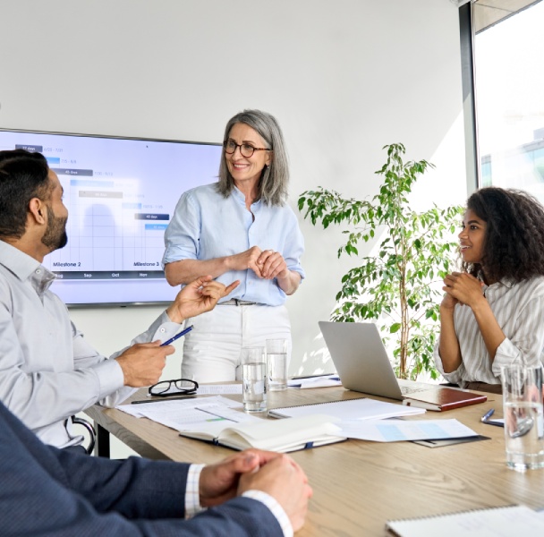 Group meeting in a bright conference room, discussing data presented on a screen, with laptops and documents on the table.