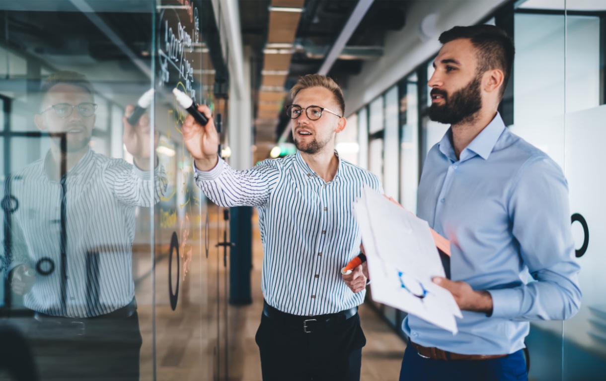 Two professionals engage in a discussion at a glass wall, one writing with markers while the other holds notes and observes.