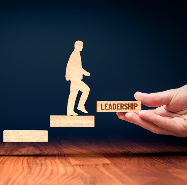 A wooden figure climbs stairs, while a hand presents a block reading "LEADERSHIP," symbolizing ascension in leadership.