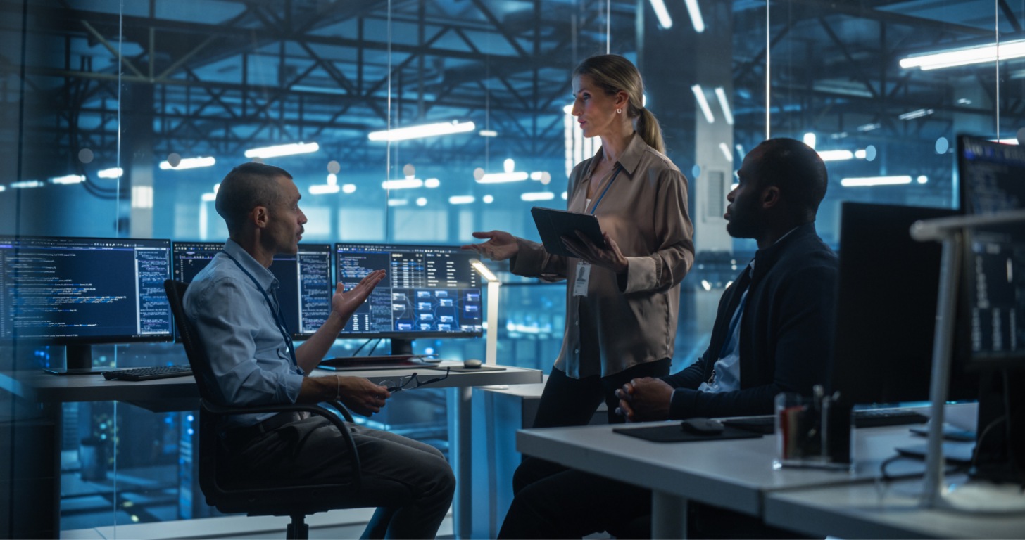 A business meeting in a modern office with multiple monitors displaying data. A woman discusses with two seated colleagues.