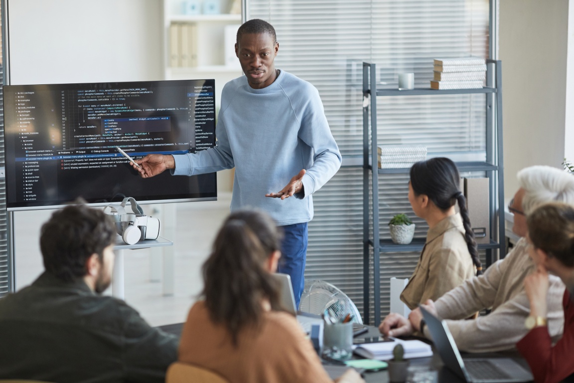 A speaker presents coding concepts on a screen to an engaged audience in a modern office setting.